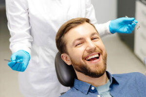 Smiling bearded man sitting in a dental chair during a check-up, with a dentist in gloves holding dental tools in the background.